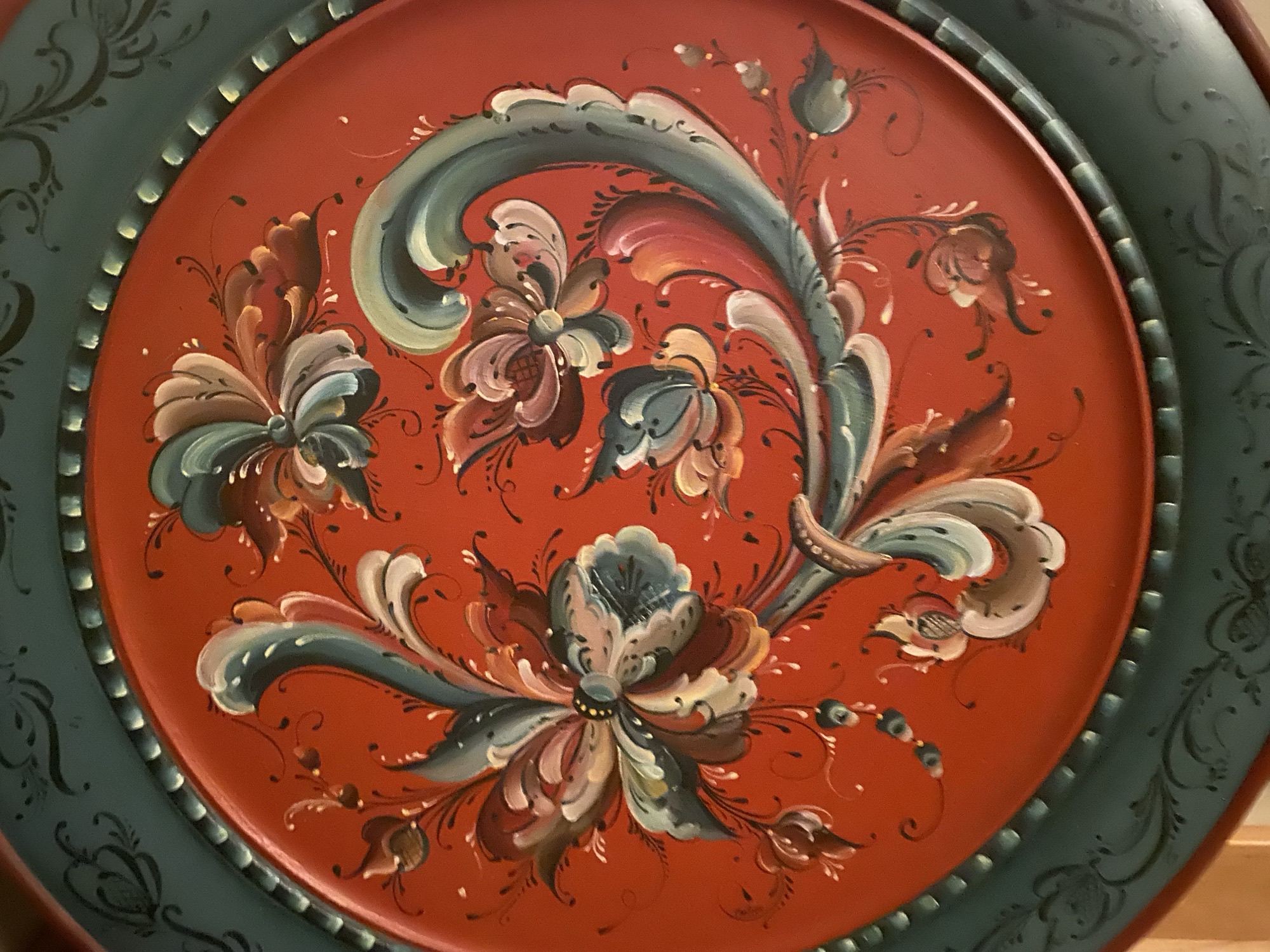 Close-up of a hand-painted plate with flowing rosemaling flowers and scrolls in slate blue, cream, and gold on a deep barn-red ground.