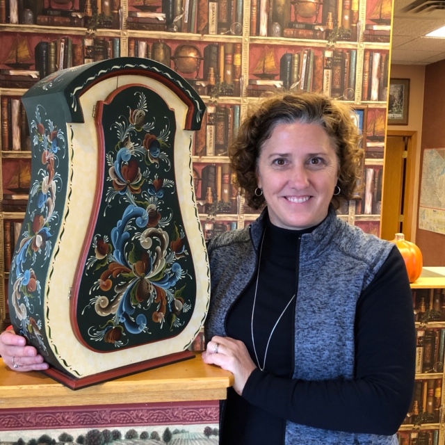 Michelle Carlson holding a large hand-painted cabinet with dark green ground and rosemaling scrollwork.